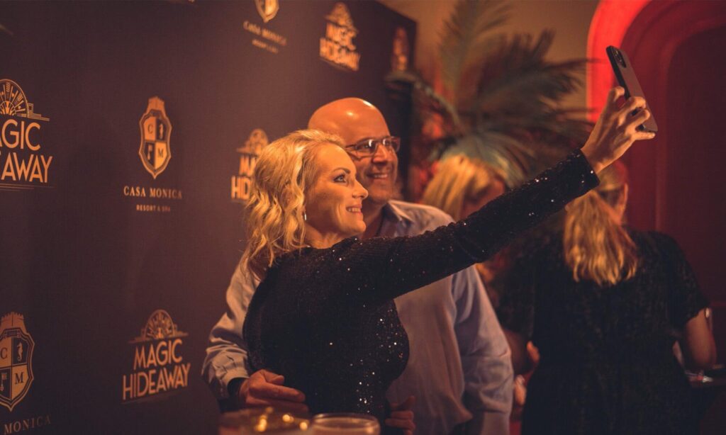 A smiling couple on a date night, taking a selfie in front of the Magic Hideaway theater photo wall