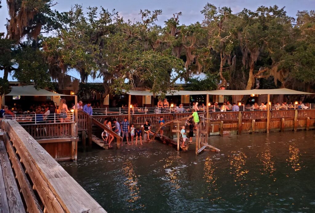 People dining at a wooden restaurant overlooking ocean at sunset and trees canopy in background