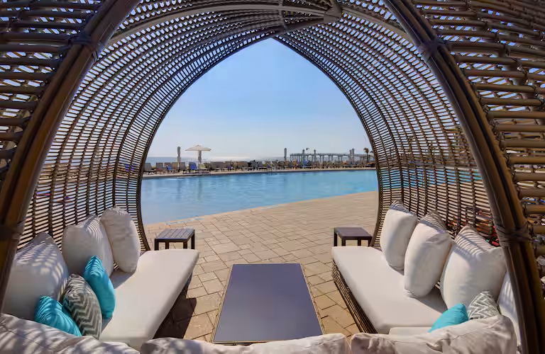 A brown cabana with white couches and teal pillows overlooking a pool and blue sky at Embassy Suites, St. Augustine, Fl