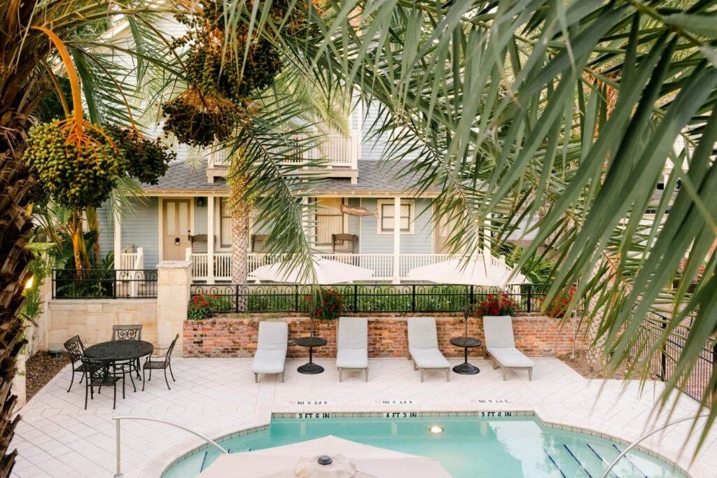 A pool, light patio, sandy colored lounge chairs with a red brick wall behind and a house at Collector Inn, St. Augustine