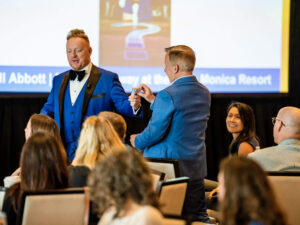 Bill Abbott in blue tuxedo and guest in blue suit choosing a card while specators watch and screen in background