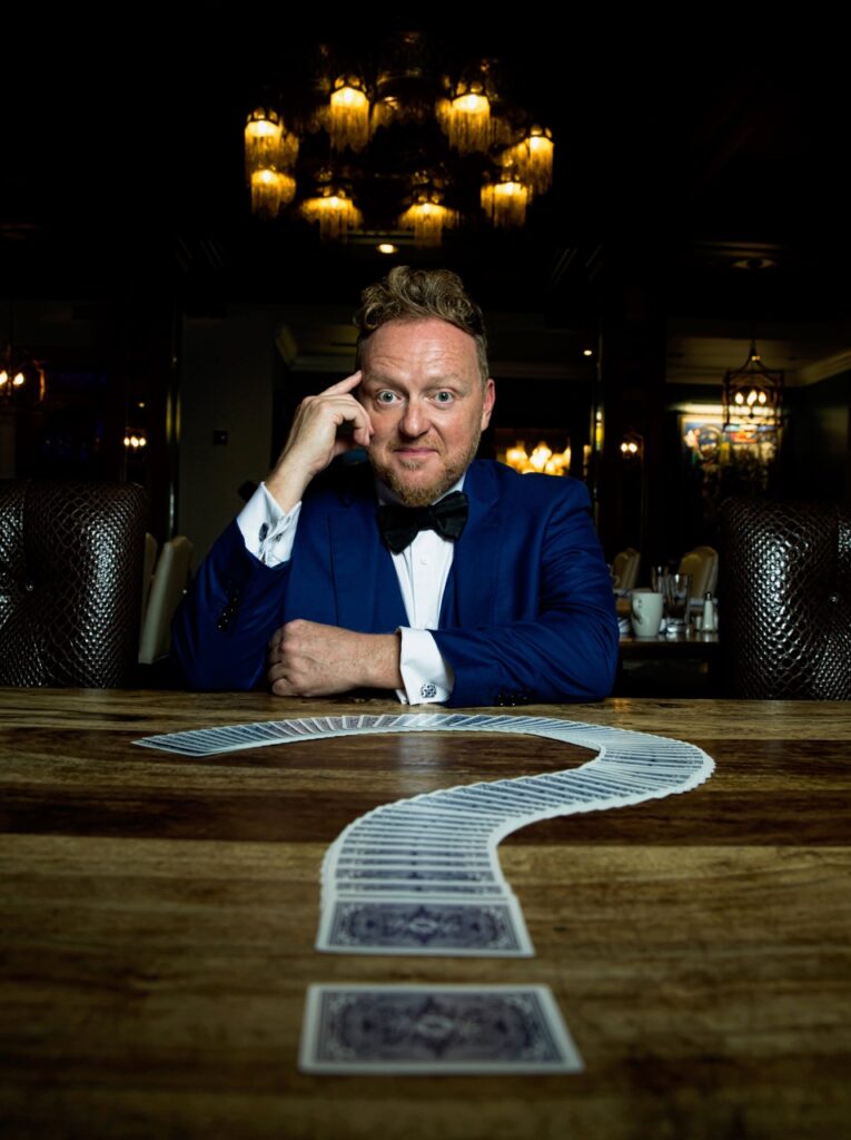 Bill Abbott magician sitting with hand on temple in blue tuxedo and question mark of cards in front on wooden table