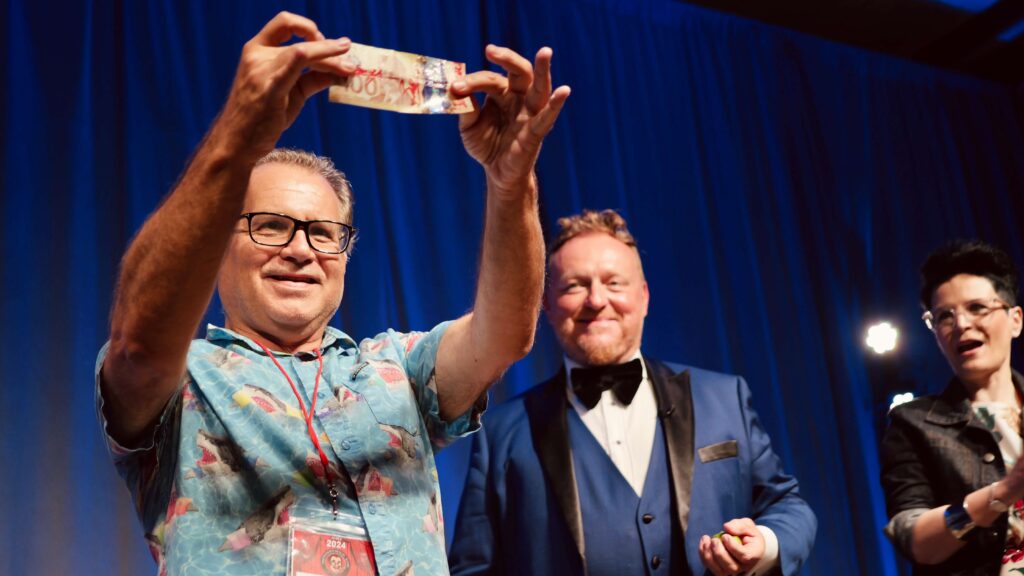 man in blue shirt holding $100 bill in air beside magician in blue tuxedo and blue curtain background
