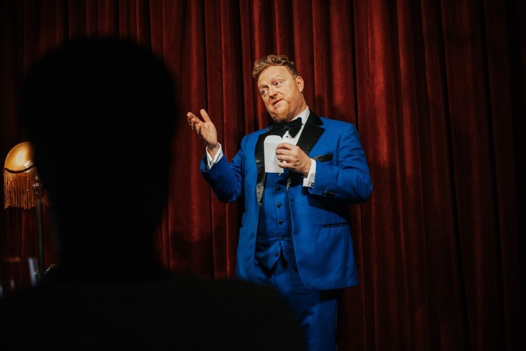 Man in blue tuxedo holding envelope with red curtain backdrop