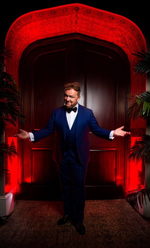 magician in blue suit with hands out wide in front of wooden door and red lighting