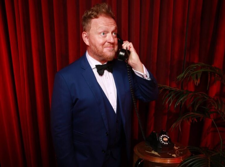 Bill Abbott magician in blue tuxedo holding phone to ear with red velvet curtains draped behind