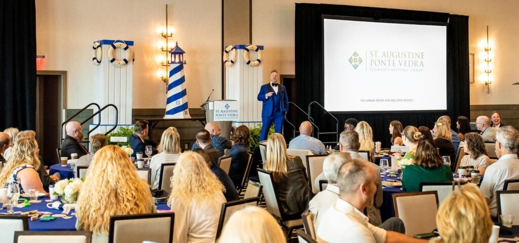 Bill Abbott in blue tuxedo performing magic for several people in crowd at conference in bright room.