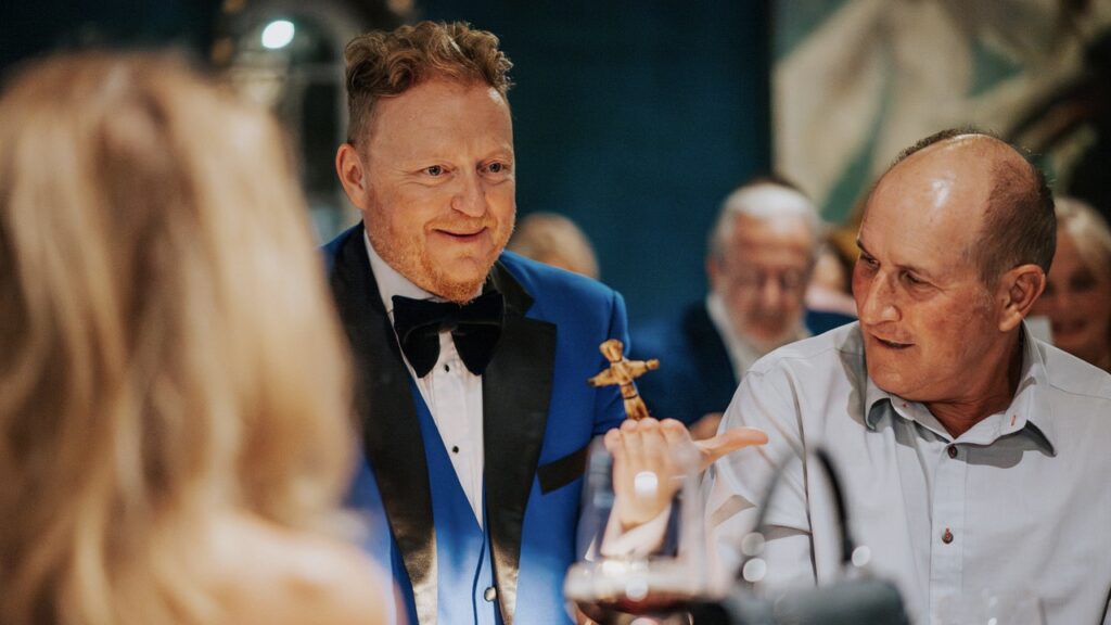 Bill Abbott in blue tuxedo and holding magic trick for guest in light blue shirt who looks at performance