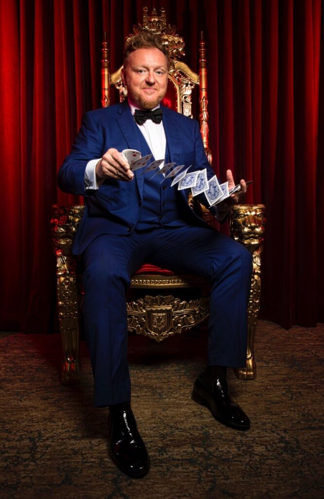 Magician in blue tuxedo sitting in tall gold chair, red curtains behind, with cards flying from one hand to another.