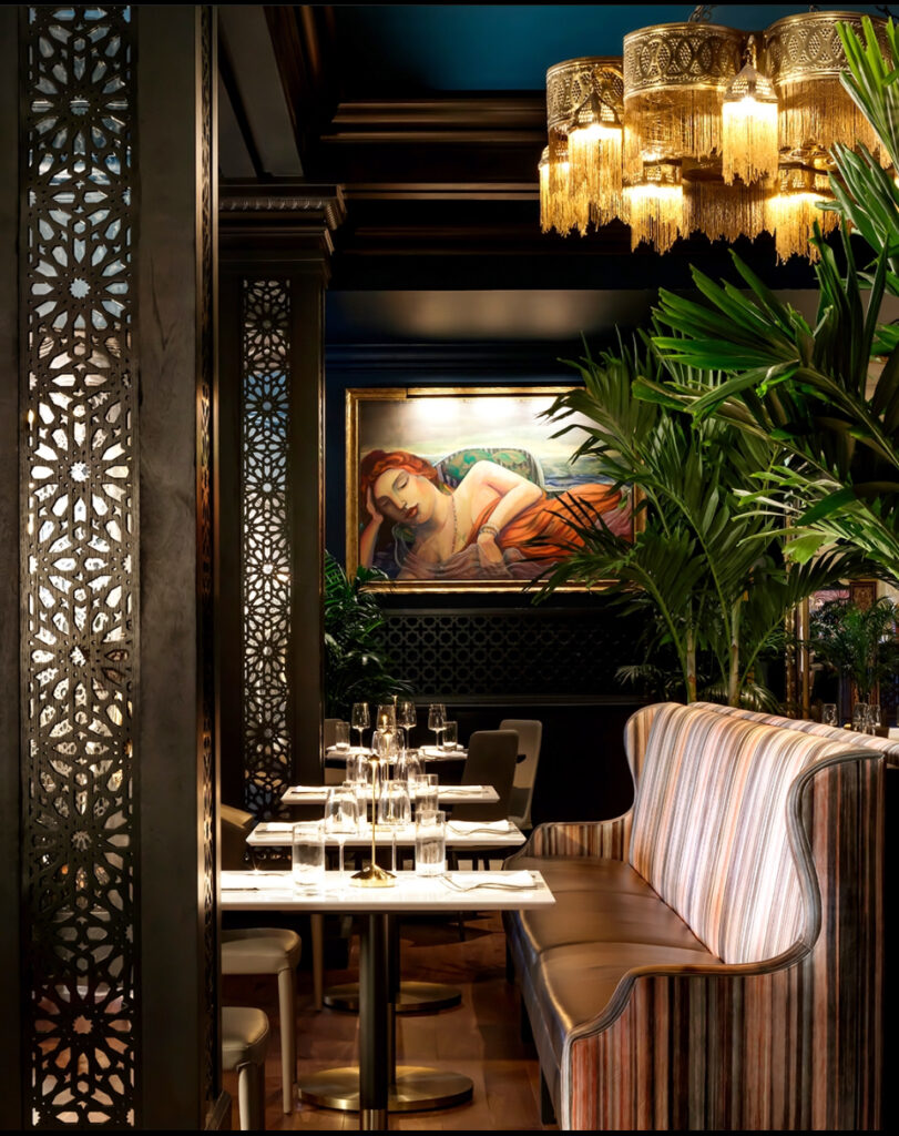 Pink booth seating, gold ceiling light, painting of a woman, black pillars, white tables with glasses, palm plants