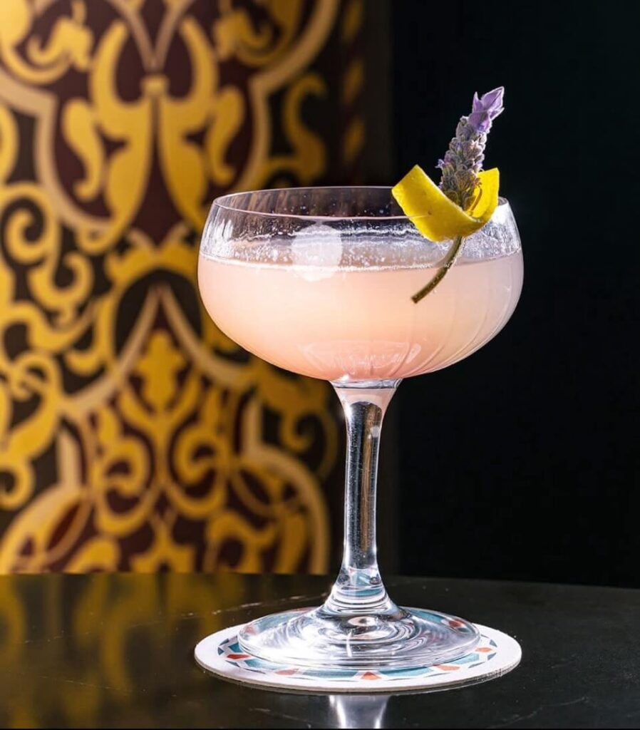 Pink cocktail in a coup glass, garnished with lavender and lemon peel with a gold and burgundy printed wallpaper in back.