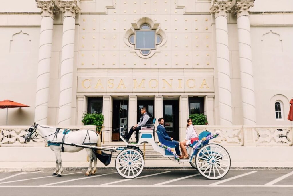White horse drawn carriage with couple sitting in back in front of light building with Casa Monica Sign