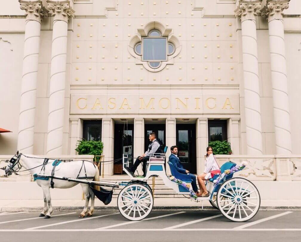 White horse drawn carriage with couple sitting in back in front of light building with Casa Monica Sign