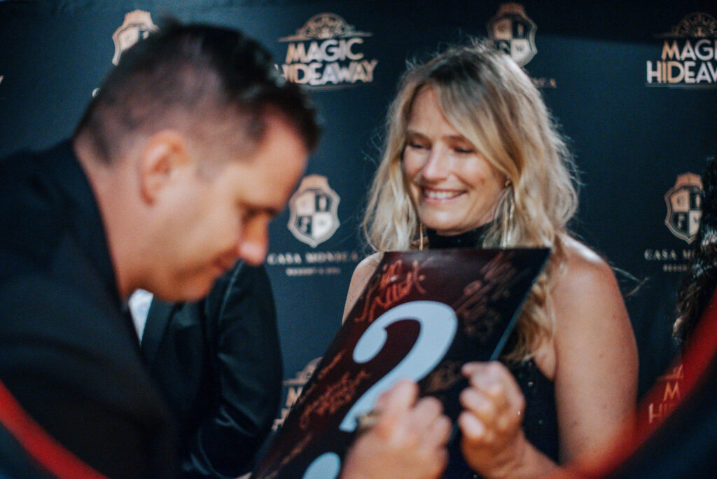 gentleman in black signing a question mark envelope and woman in black dress against Magic Hideaway logo photo wall