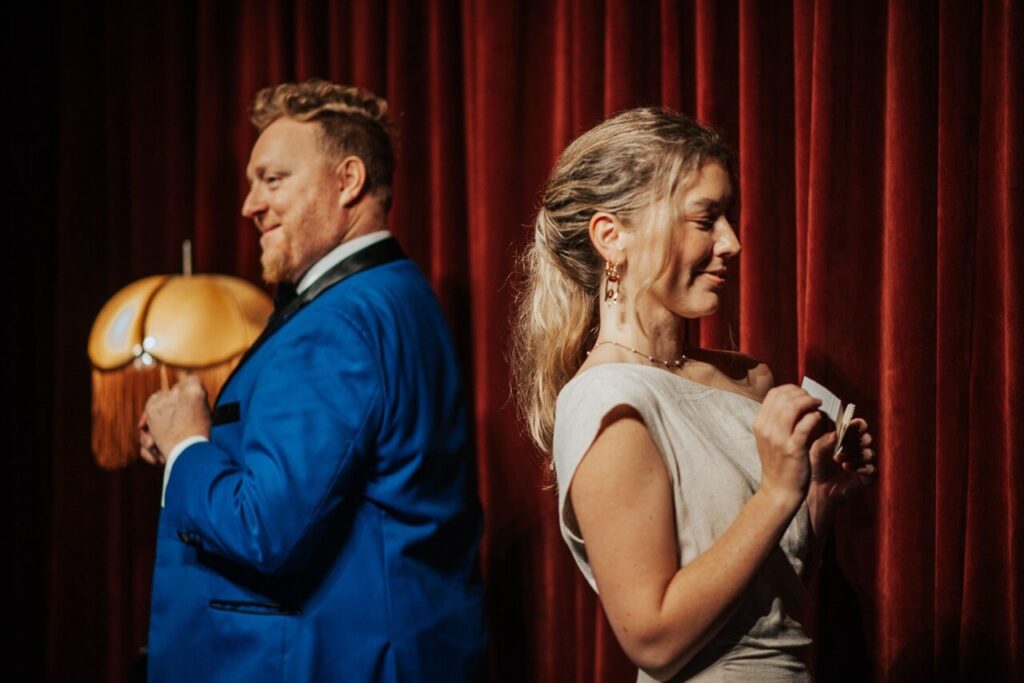 Man in blue suit and woman in tan dress hold paper with red curtains and lamp