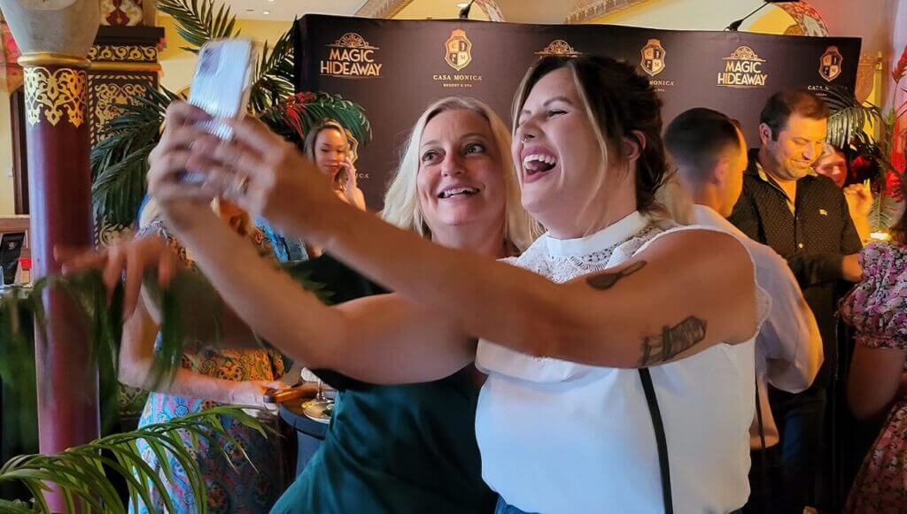 Two woman smile for selfie with guests in background in front of Magic Hideaway logo photo wall